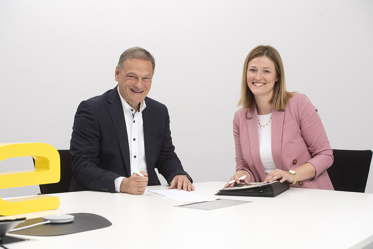 Ein Mitarbeiter und eine Mitarbeiterin von e-mobil BW sitzen an einem Tisch und schauen in die Kamera. Sie befinden sich in einem Bewerbungsgespräch, während der Mitarbeiter gerade einen Vertrag unterzeichnet.