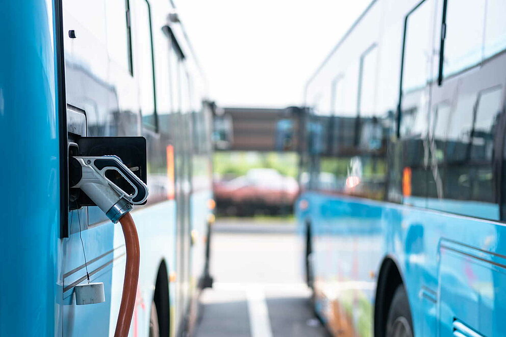 Zwei nebeneinanderstehende E-Busse beim Stromladen auf einem Parkplatz.