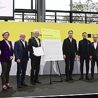 Eine Ansammlung von Personen auf dem Podium des Strategiedialogs Automobilwirtschaft BW. Ministerpräsident Winfried Kretschmann hält ein wichtiges Dokument in der Hand.