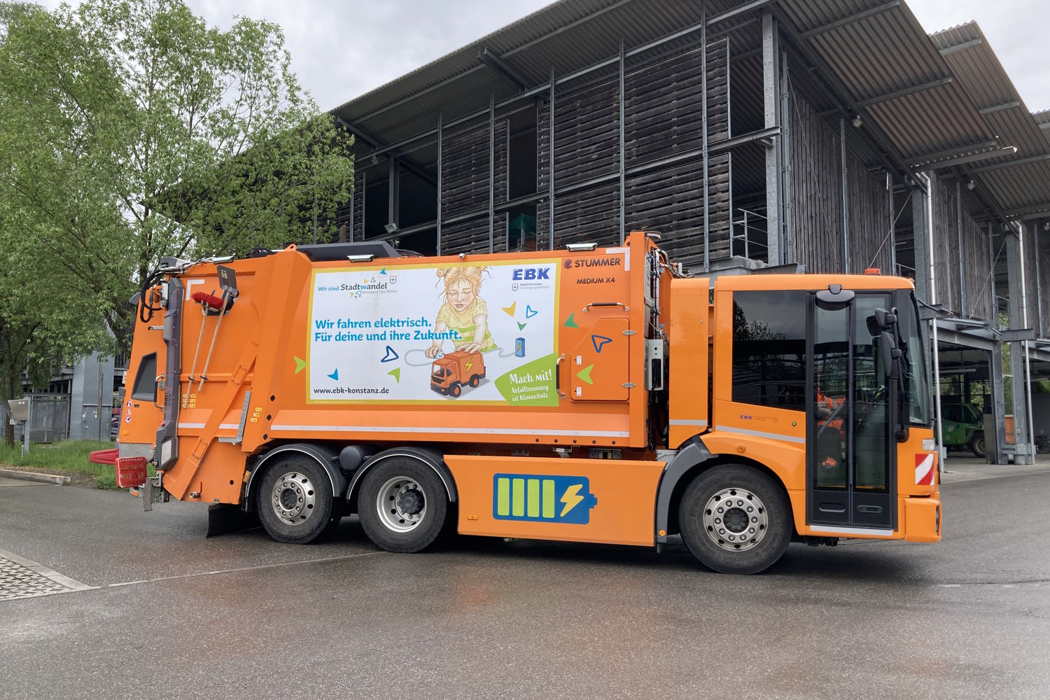 Ein orangenes E-Müllfahrzeug vor einem Gebäude.