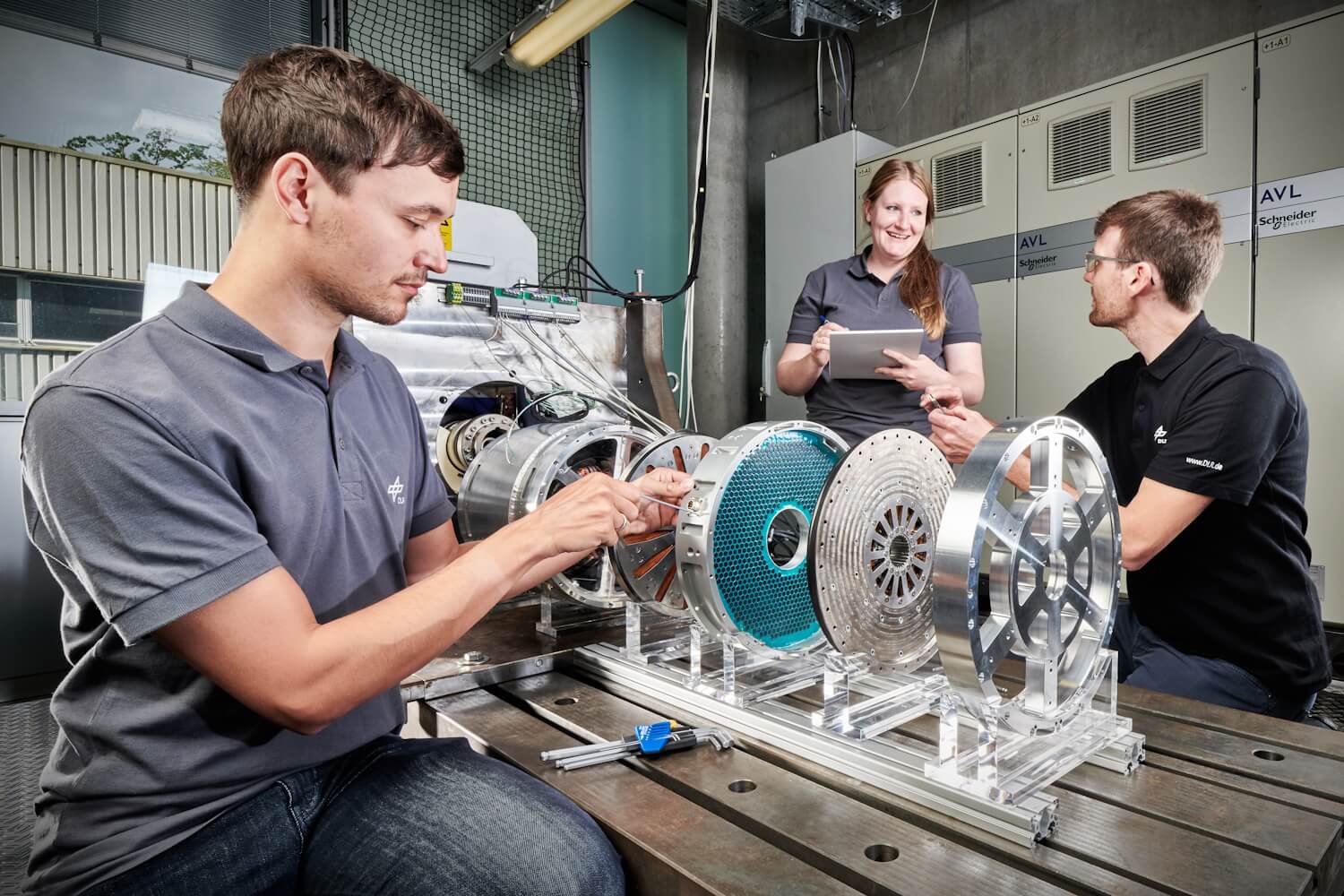 Das Bild zeigt drei Personen, die um eine Werkbank stehen und eine elektrische Maschine bedienen..